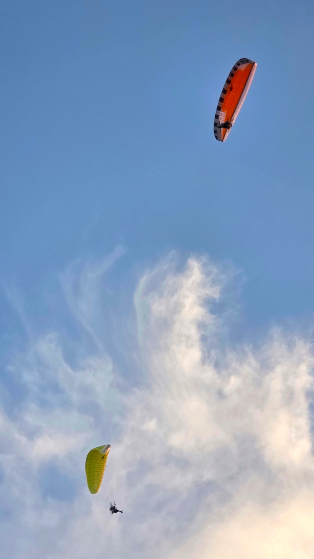 Two paramotor pilots flying high under orange and yellow wings